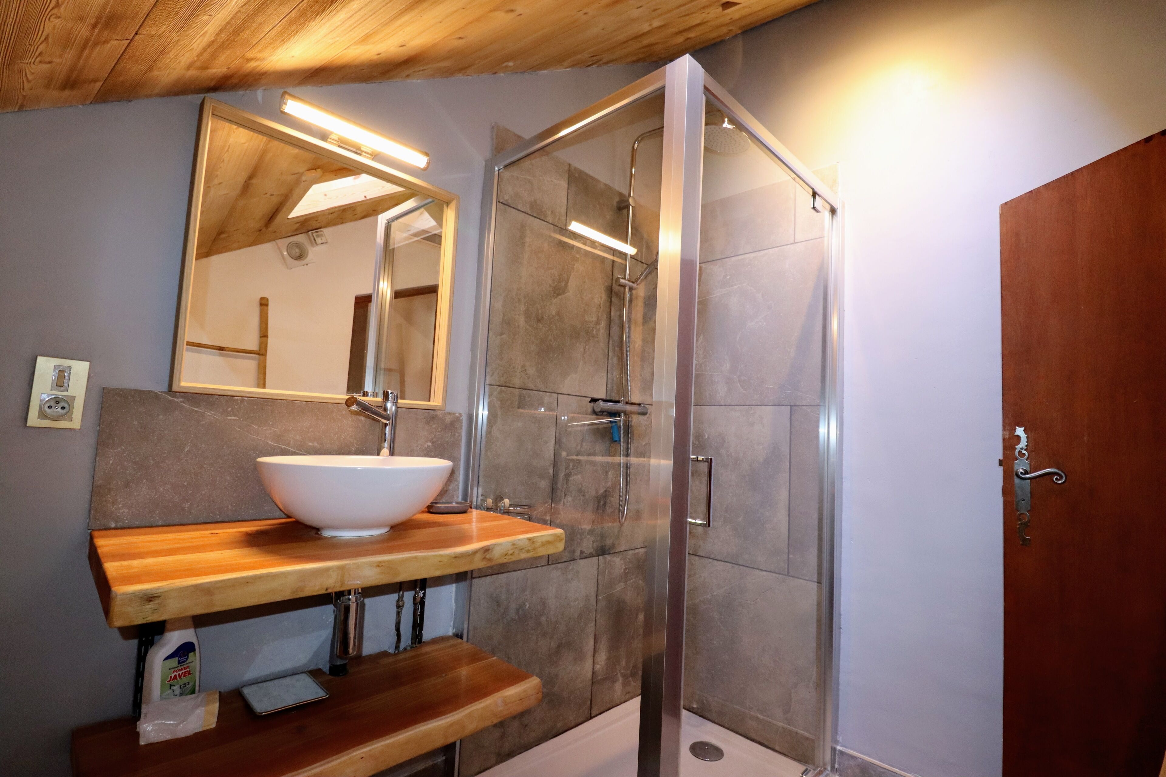 A bathroom with a wooden vanity, a white vessel sink, a glass shower enclosure, and a wooden door.