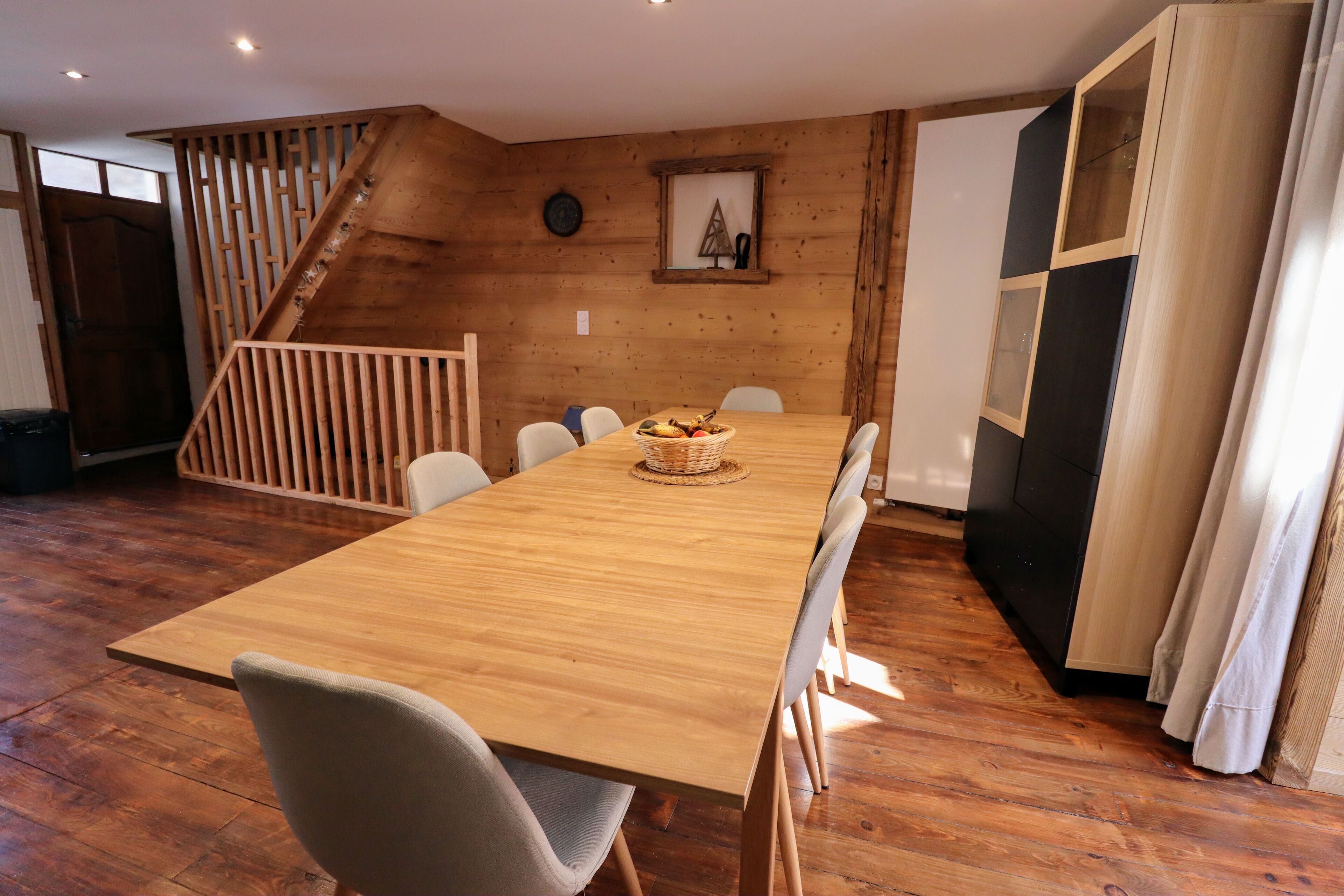 Une table à manger en bois, des chaises, un panier de fruits et une horloge au mur.