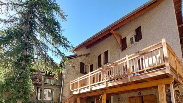 A rustic two-story building with a wooden balcony, a stone facade, and a well-maintained garden with a tree and flower beds.