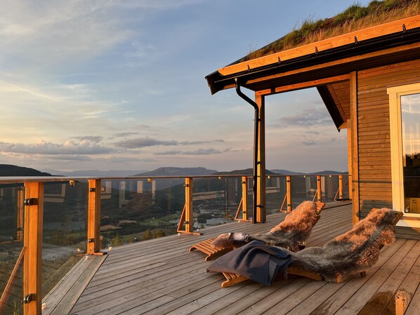 Une terrasse en bois avec deux chaises longues, dont une est recouverte d’une couverture et sur laquelle se trouve un smartphone, et une grande fenêtre donnant sur une vallée.