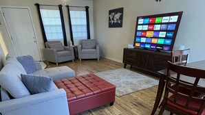 A living room with a grey sofa, a red ottoman, a wooden dining table with a red chair, and a wall-mounted TV displaying various streaming services. - Cute Townhouse in Cherokee Village where adventures await! (Cherokee Village)