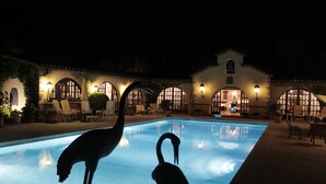 A swimming pool with two flamingos, surrounded by lounge chairs and a building with arched windows and outdoor lighting. - Masia Mimosa (Darnius)