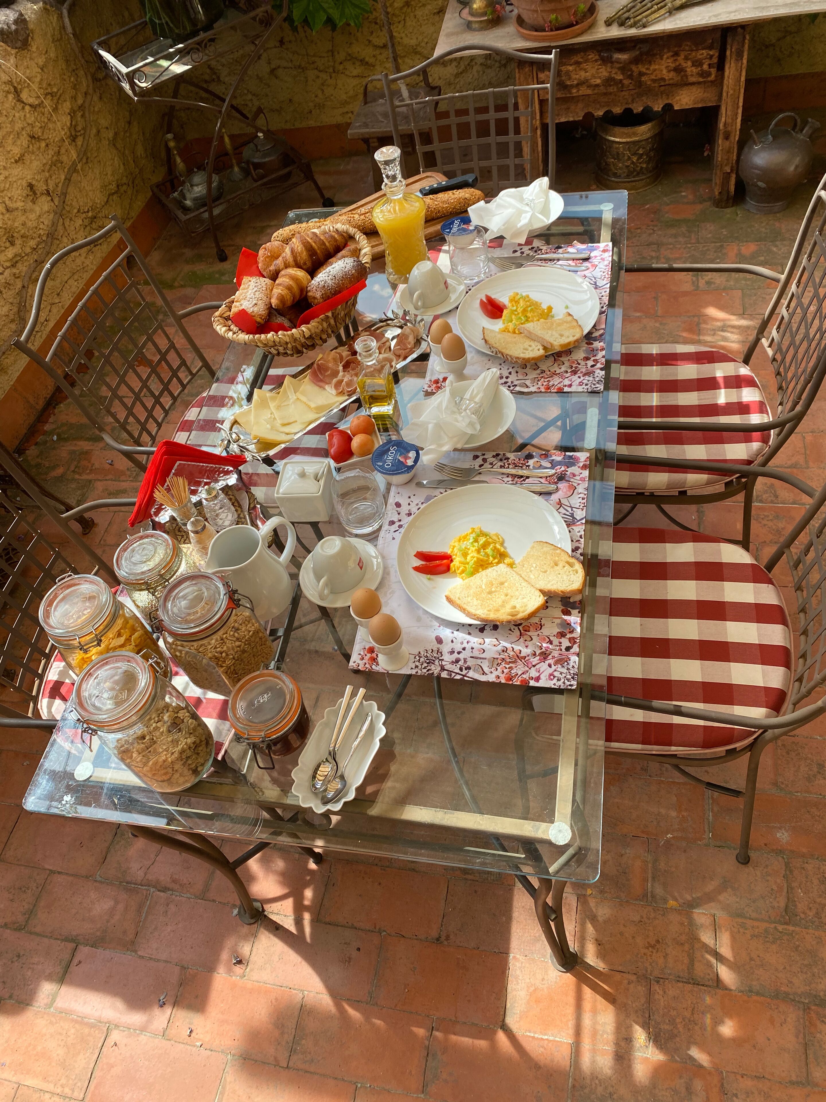 Mesa de desayuno al aire libre con diferentes alimentos, utensilios y una mesa de vidrio.