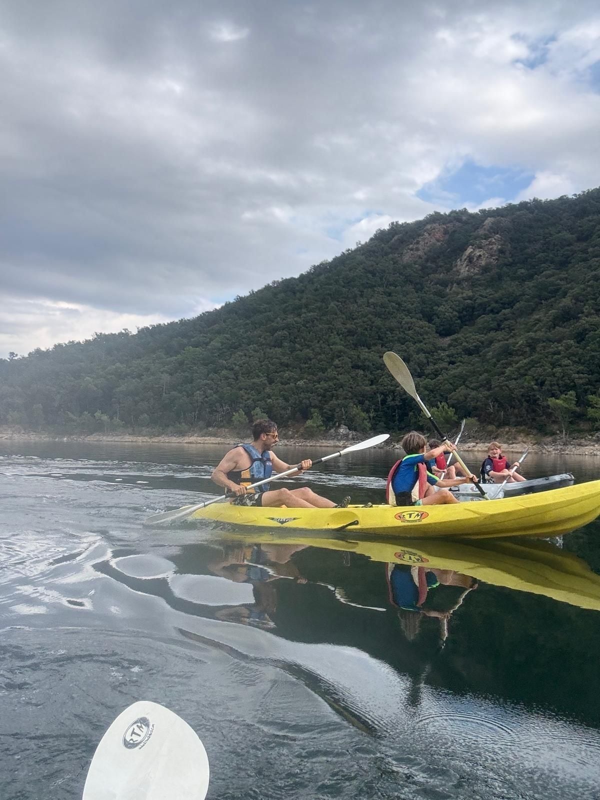 A group of people in a yellow kayak on a calm body of water with a forested hill in the background.