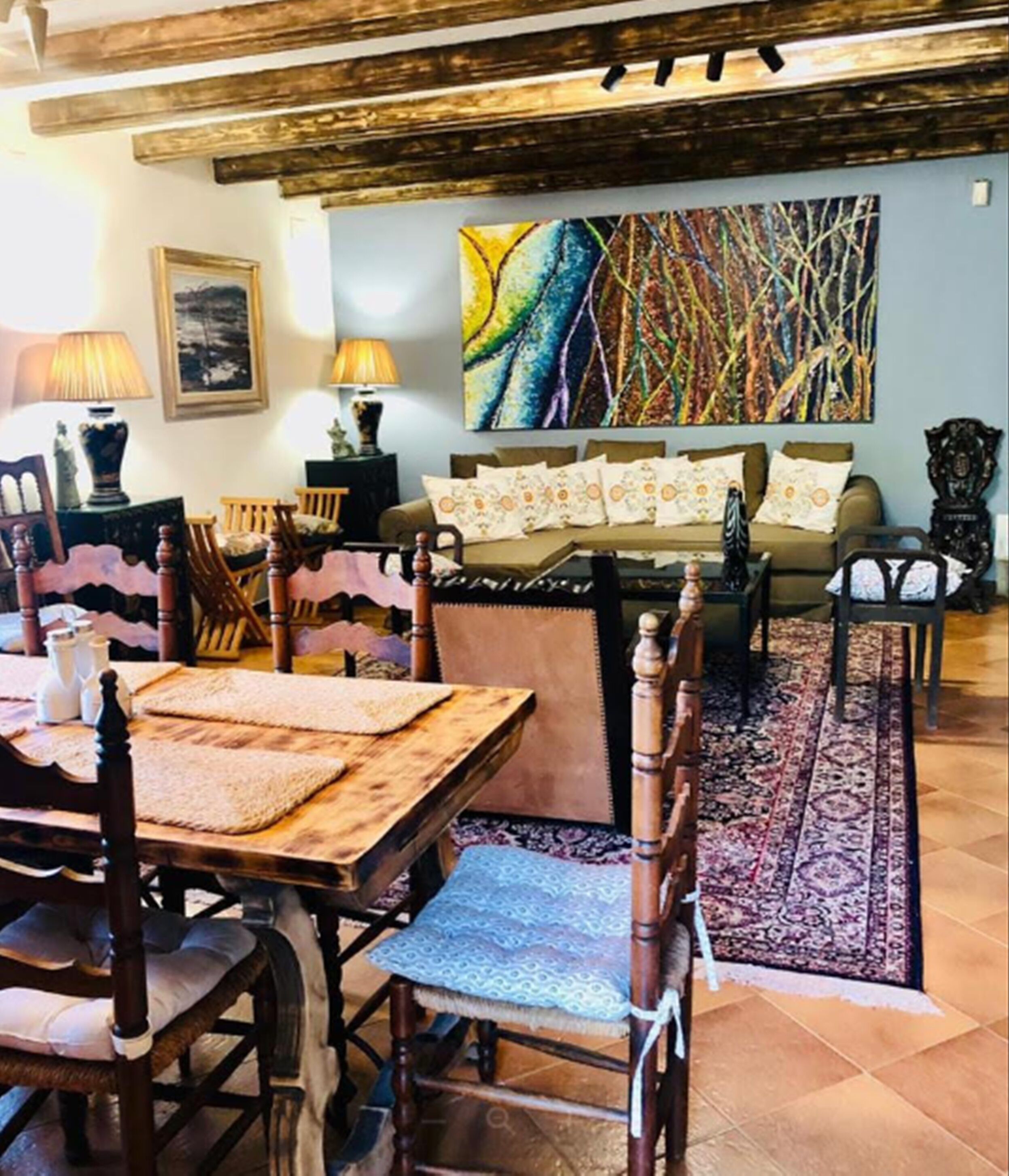 Un área de comedor con una mesa y sillas de madera, un conjunto de sofás y un cuadro abstracto y colorido en la pared.