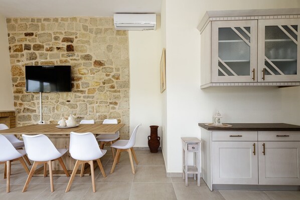 A dining area with a stone wall, a flat-screen TV, white chairs, a wooden table, and white cabinets.