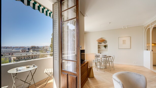 A balcony with a view of buildings, a dining area with chairs and a table, and a wooden floor.
