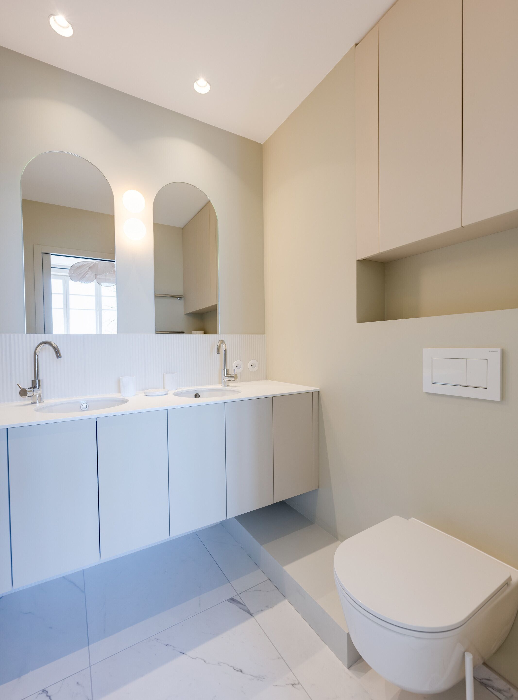 Et moderne badeværelse med en hvid vask, to haner, et spejl og et toilet.