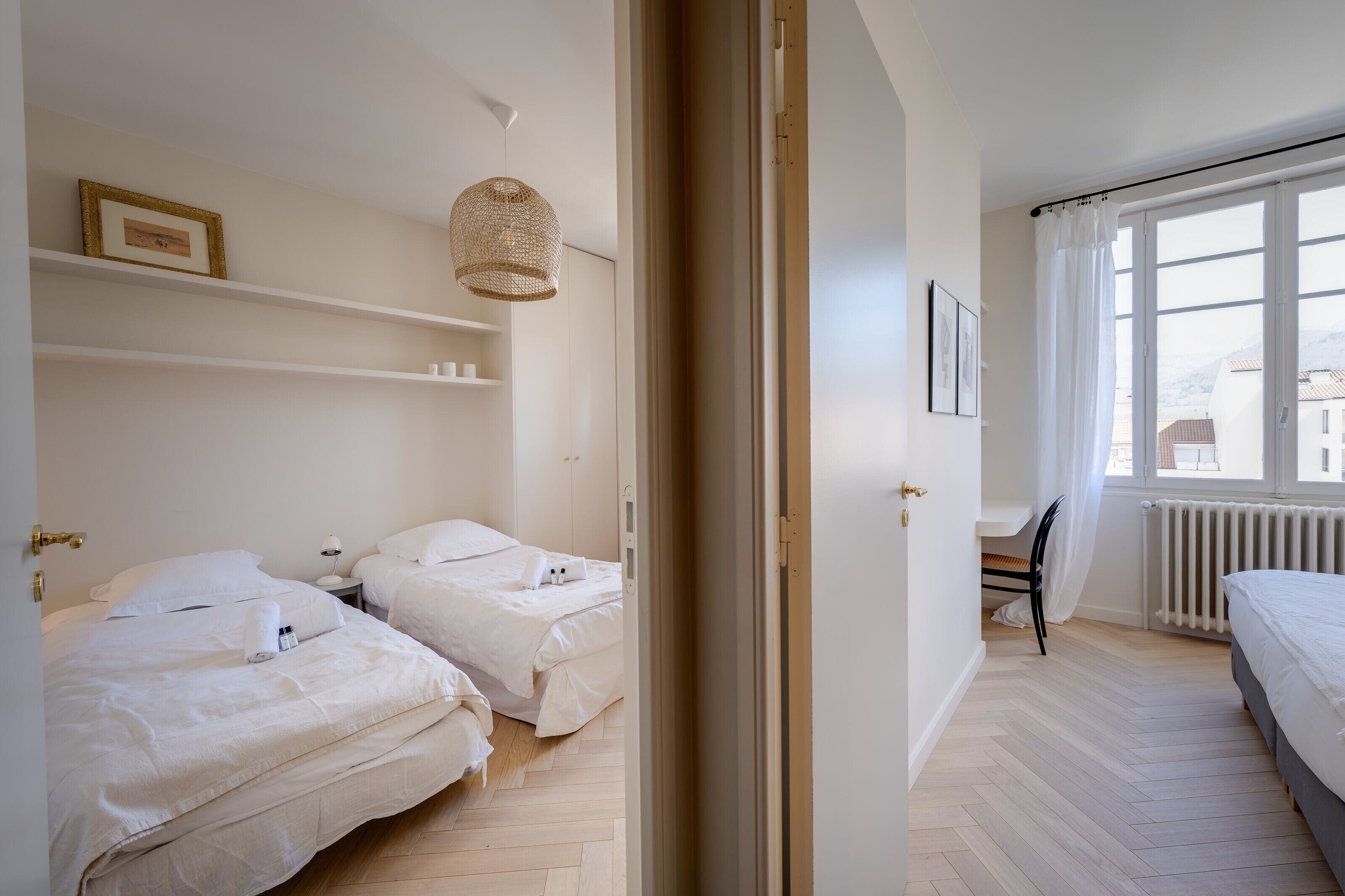 Hotellihuone, jossa on kaksi sänkyä, rottinkinen valaisin, patteri ja suuret ikkunat, joista avautuu näkymä kaupunkiin.