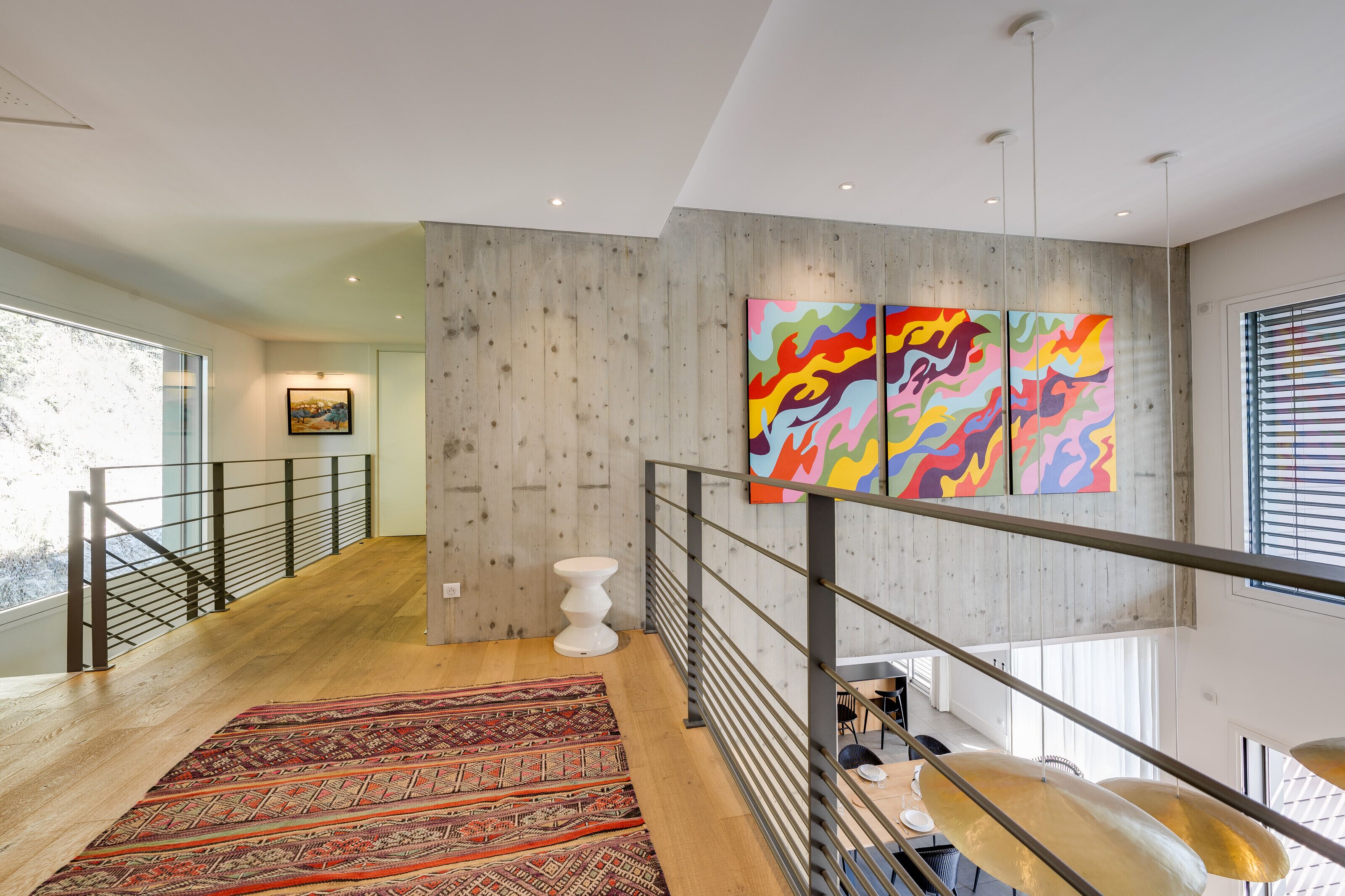Un intérieur moderne avec un plancher en bois, un tapis à motifs, un escalier avec des rampes en métal et une peinture abstraite aux couleurs vives accrochée au mur.