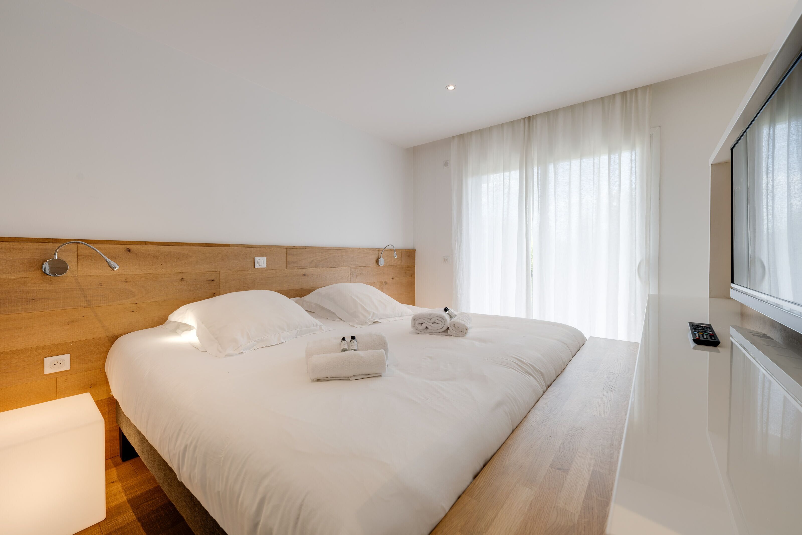 Una habitación de hotel moderna con cabecera de madera, una cama grande con ropa blanca, una mesita de noche y una ventana con cortinas.