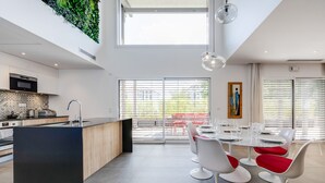 A modern kitchen with a central island, large windows, and a dining area with white chairs and red cushions.