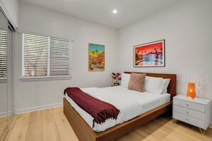 A modern bedroom with a wooden bed, white bedding, a red throw blanket, a nightstand with a lamp, and two paintings on the wall.