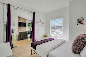 A modern bedroom with a bed, a TV mounted on the wall, and a window with blinds.
