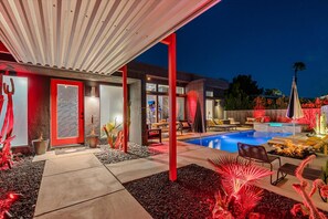 A modern outdoor pool area with a covered seating area, a red door, and a well-lit entrance.