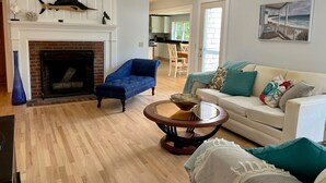 Una sala de estar con chimenea, un sillón azul, una mesa de centro redonda y un sofá beige con cojines estampados.