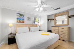 Ein Schlafzimmer mit einem Bett, zwei Nachttischen inklusive Lampen, einem Deckenventilator, einer hölzernen Kommode, einem großen Spiegel und einem Wandbild mit Strandmotiv.