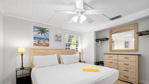 Un dormitorio con cama, dos mesitas de noche con lámparas, un ventilador de techo, un aparador de madera, un espejo grande y un cuadro con motivos playeros.