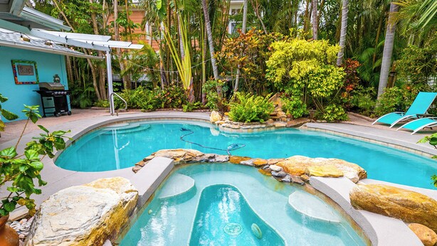 A swimming pool with a hot tub, surrounded by rocks and plants, and a barbecue area. - Pool Party South · 3BR · Pool & Spa · Updated Jan 2025 (Holmes Beach)