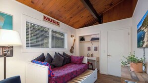 A cozy room with a bed, a desk, a guitar, and a window with blinds.