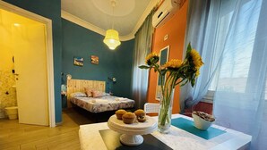 Un dormitorio moderno con suelo de madera, una cama con colcha estampada de flores, una mesita de noche con un jarrón de girasoles y una ventana con cortinas de gasa.