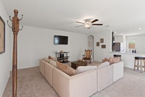 A living room with a large beige sectional sofa, a wooden floor lamp, a ceiling fan, a flat-screen TV, and a small table with decorative items.