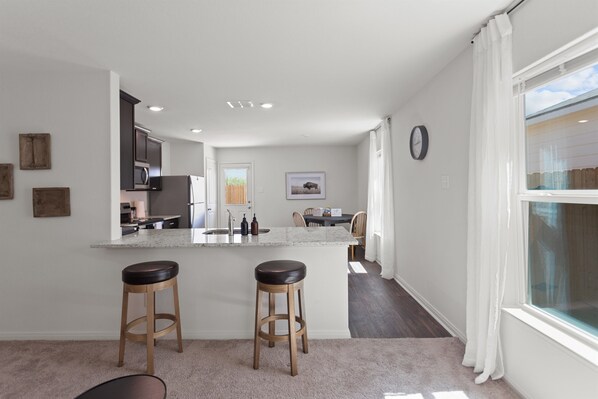 A modern kitchen with a central island, two bar stools, and a dining area with a table and chairs.