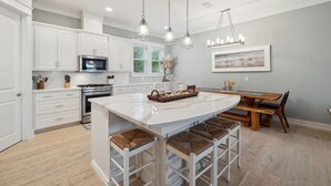 Une cuisine moderne avec des meubles blancs, un îlot central doté de tabourets de bar, un éclairage suspendu et un coin repas avec une table et des bancs en bois.