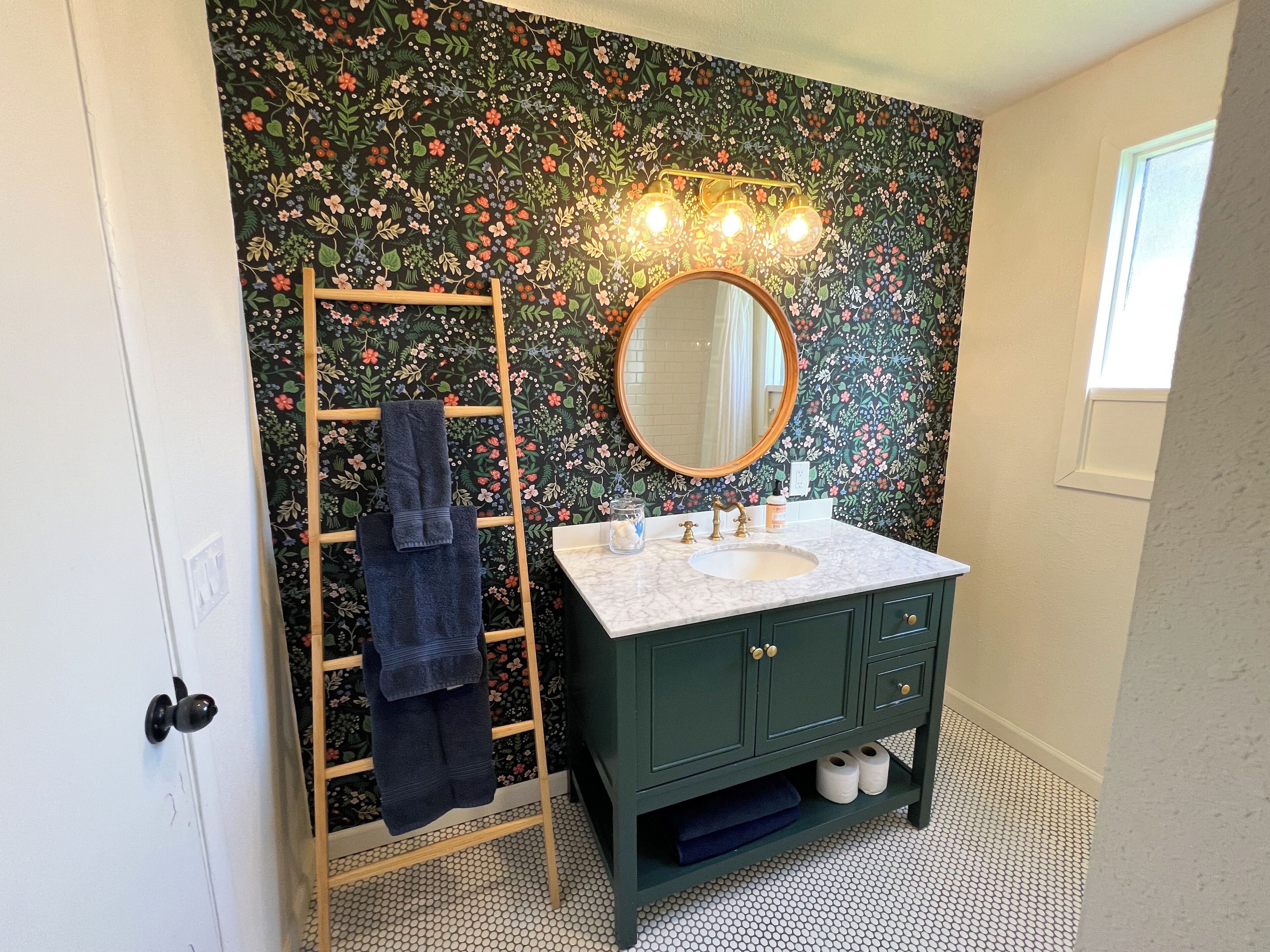 A bathroom with a floral wallpaper, a wooden ladder with a towel, a round mirror, and a sink with a marble countertop.