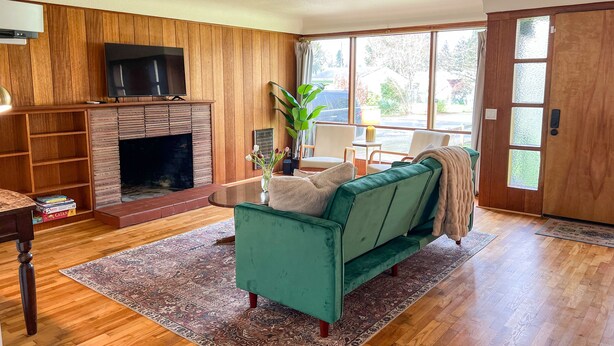 A living room with a fireplace, a green sofa, a TV, and a large window. - Whimsical Midcentury Home with Game Room (Springfield)