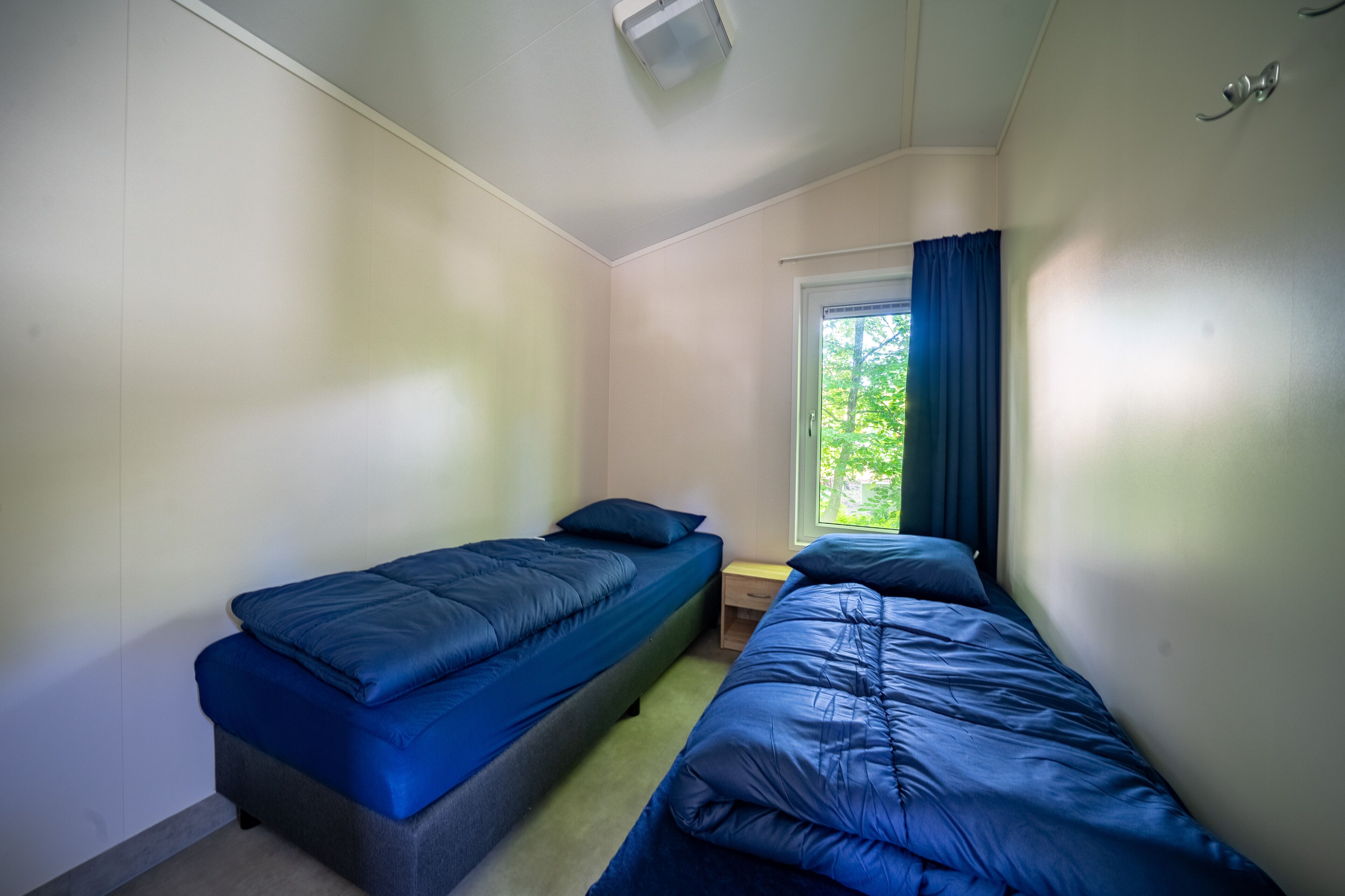 A small, single-room accommodation with two single beds, a window with curtains, and a ceiling light.