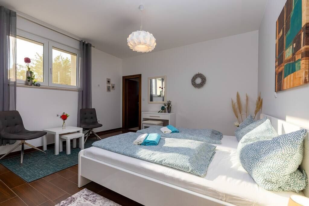 A modern bedroom with a bed, two chairs, a small table, a mirror, and a window with curtains.