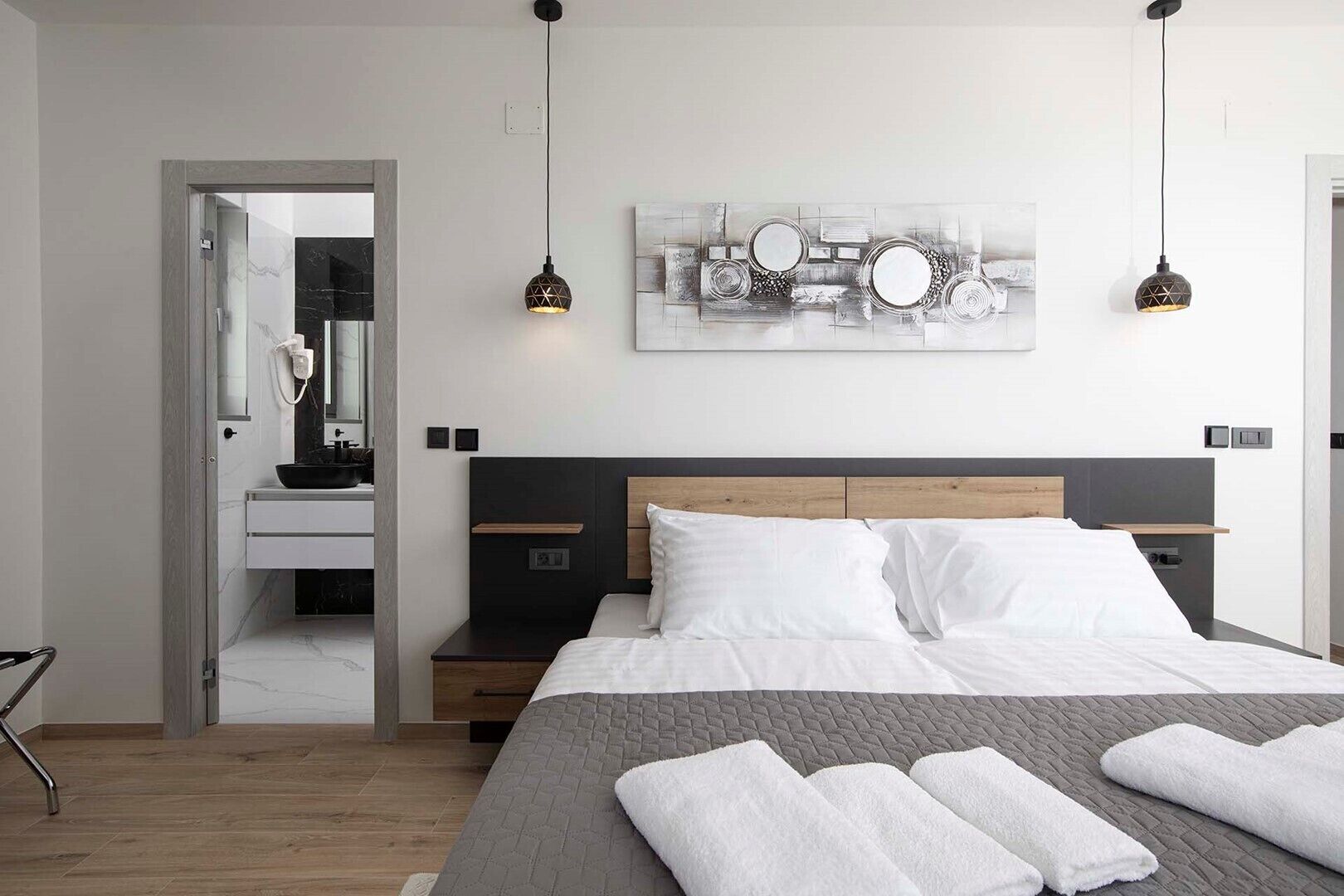 A modern bedroom with a large bed, bedside tables, a nightstand, a bathroom with a mirror, and a wall art piece.