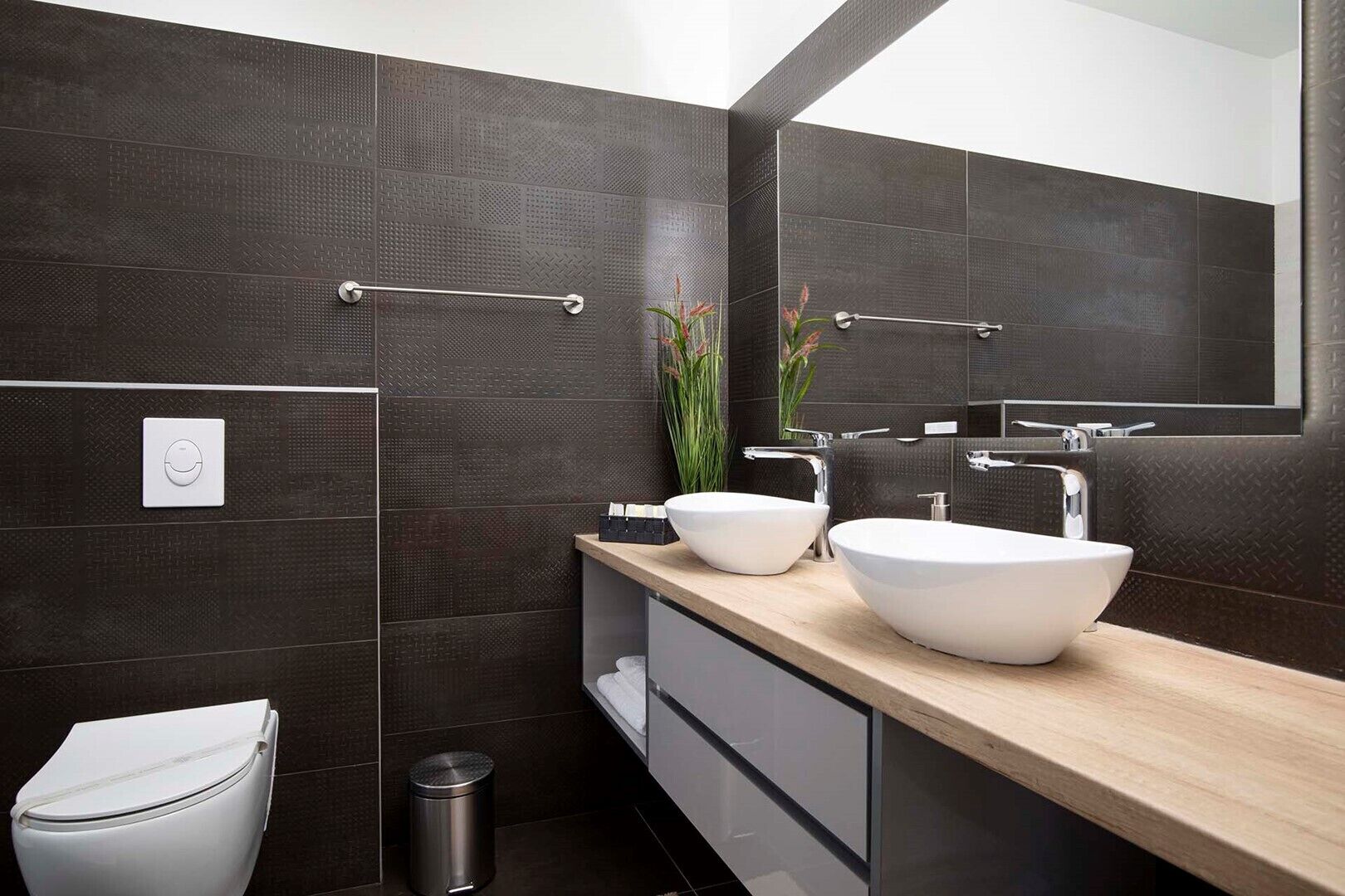Un baño moderno con paredes de azulejos oscuros, dos lavamanos blancos tipo recipiente, una encimera de madera, un espejo y un inodoro.