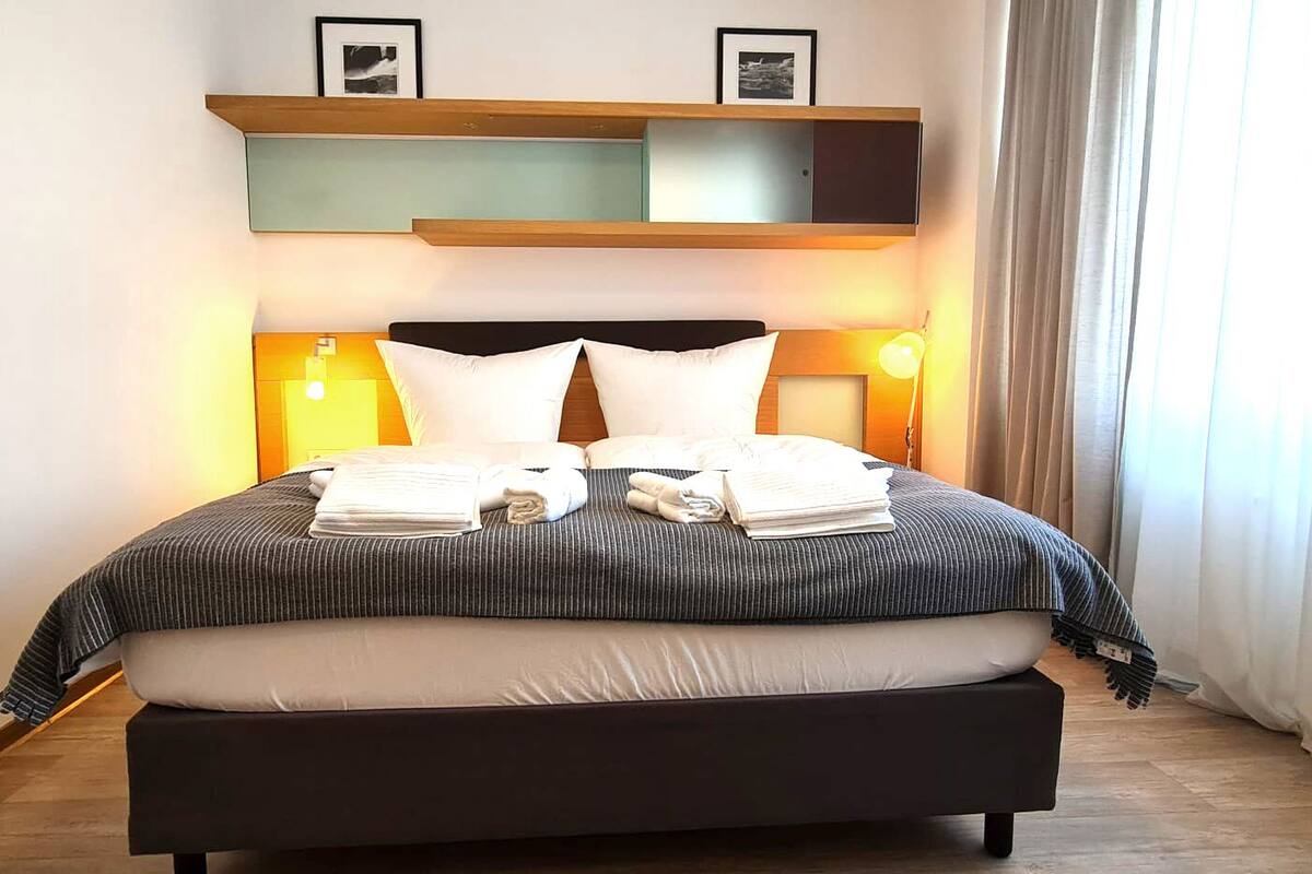 Ein ordentlich bezogenes Bett mit gestreifter Bettdecke und zwei Kissen, ein Nachttisch mit Lampe und ein oberhalb des Bettes angebrachtes Regal mit gerahmten Bildern.
