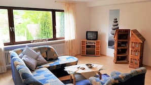 Een woonkamer met een blauwe bank, een houten boekenkast, een televisie en een raam met gordijnen.