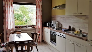 Een kleine keuken met een eettafel, stoelen, een raam met gordijnen en eenvoudige apparaten.