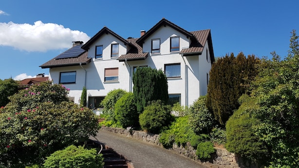 Een tweelaags huis met een schuine kap, meerdere ramen en een tuin met verschillende planten en bloemen.