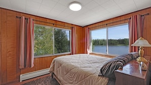 Un dormitorio con una cama grande, paredes de madera y vistas a un lago a través de dos ventanas.