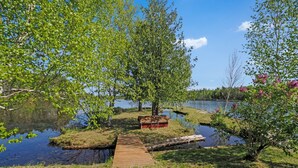 Un muelle de madera que lleva a una pequeña isla con un cobertizo rojo, rodeado de exuberante vegetación y un lago tranquilo.