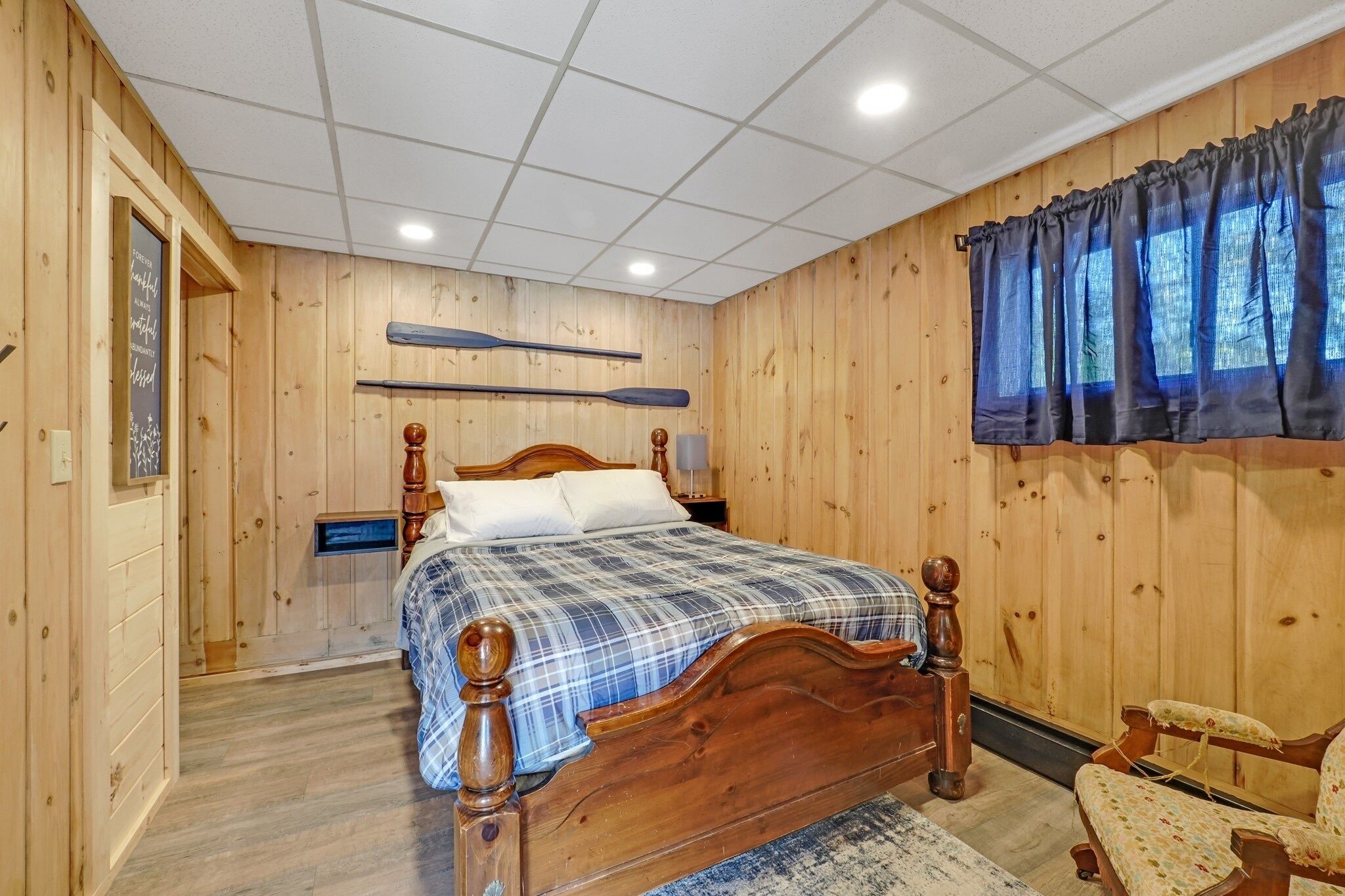 Een slaapkamer met houten lambrisering, een houten bed, nachtkastje, stoel en raam met gordijnen.