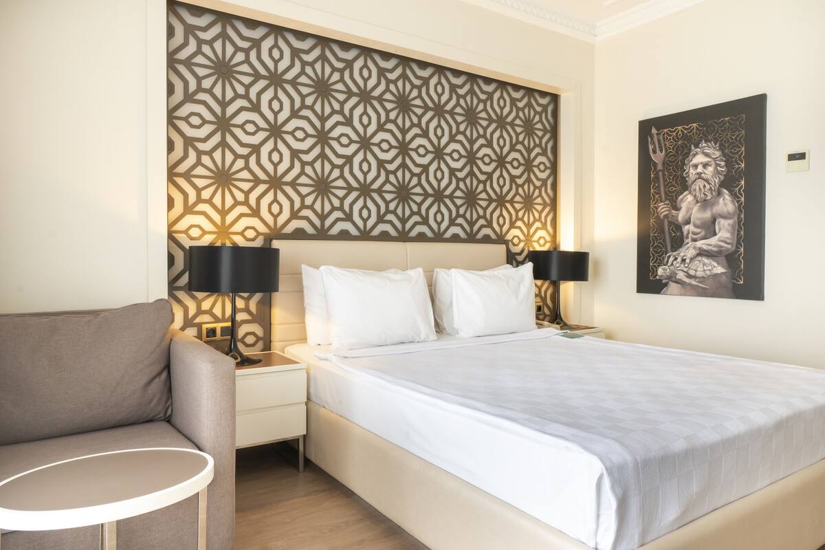 A neatly arranged hotel room with a patterned headboard, a bed with white linens, a grey sofa, and a small round table.