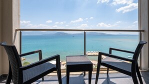 Un balcon avec deux fauteuils en osier et une petite table, d’où l’on peut admirer une mer bleue et limpide ainsi que des montagnes lointaines.
