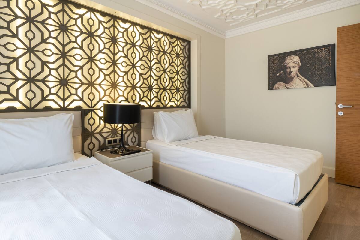 A hotel room with two beds, a decorative wall panel, a nightstand with a lamp, and a framed artwork on the wall.