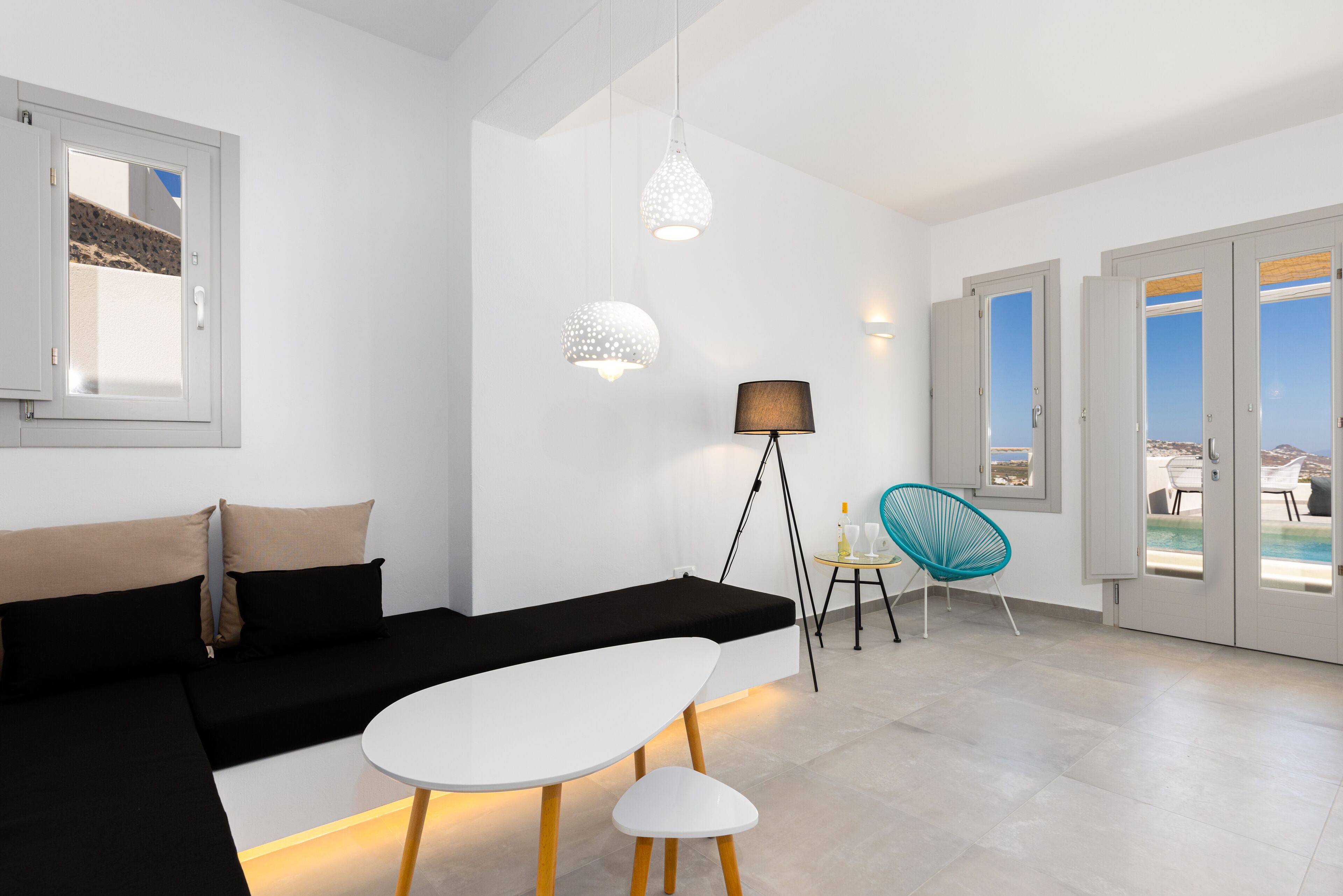 Una sala moderna con sofá negro, mesa blanca y silla verde azulada. Hay lámparas colgantes, una lámpara de pie y vistas al exterior a través de puertas de cristal.