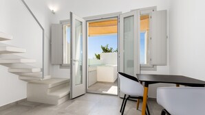 A modern interior with a staircase leading to a balcony, a dining area with a table and chairs, and a view of the sky and greenery outside. - SERAPIAS SUITES (Santorini)
