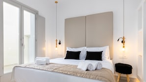 A modern hotel room with a large bed, a small stool, and a view through a sliding door. - SERAPIAS SUITES (Santorini)