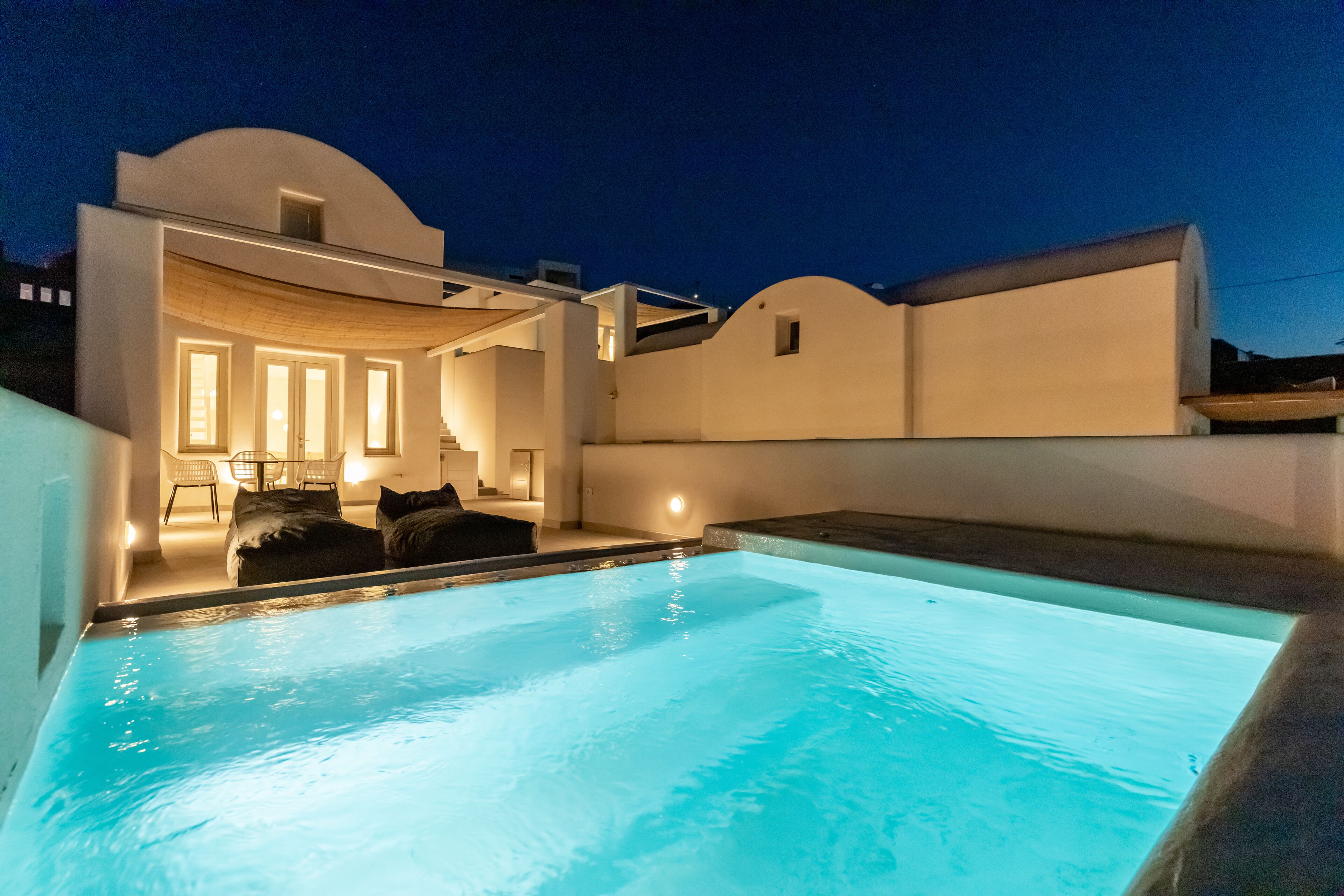 A modern pool area with a clear pool, a lounge with a sofa and chairs, and a white building with a curved roof.