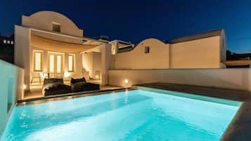A modern pool area with a clear pool, a lounge with a sofa and chairs, and a white building with a curved roof.