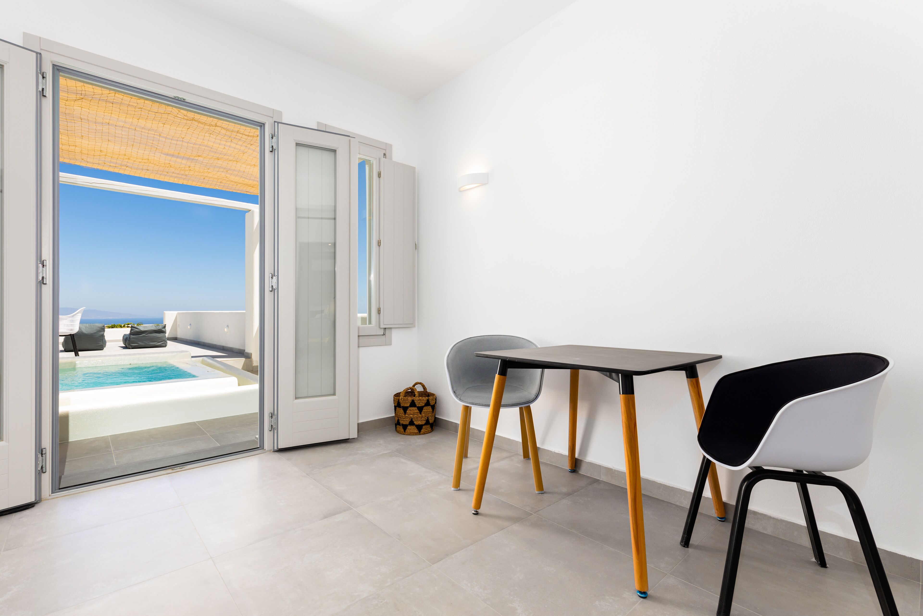 Een moderne kamer met een schuifdeur naar een balkon met een zwembad, een tafel met stoelen en een klein geweven mandje op de vloer.